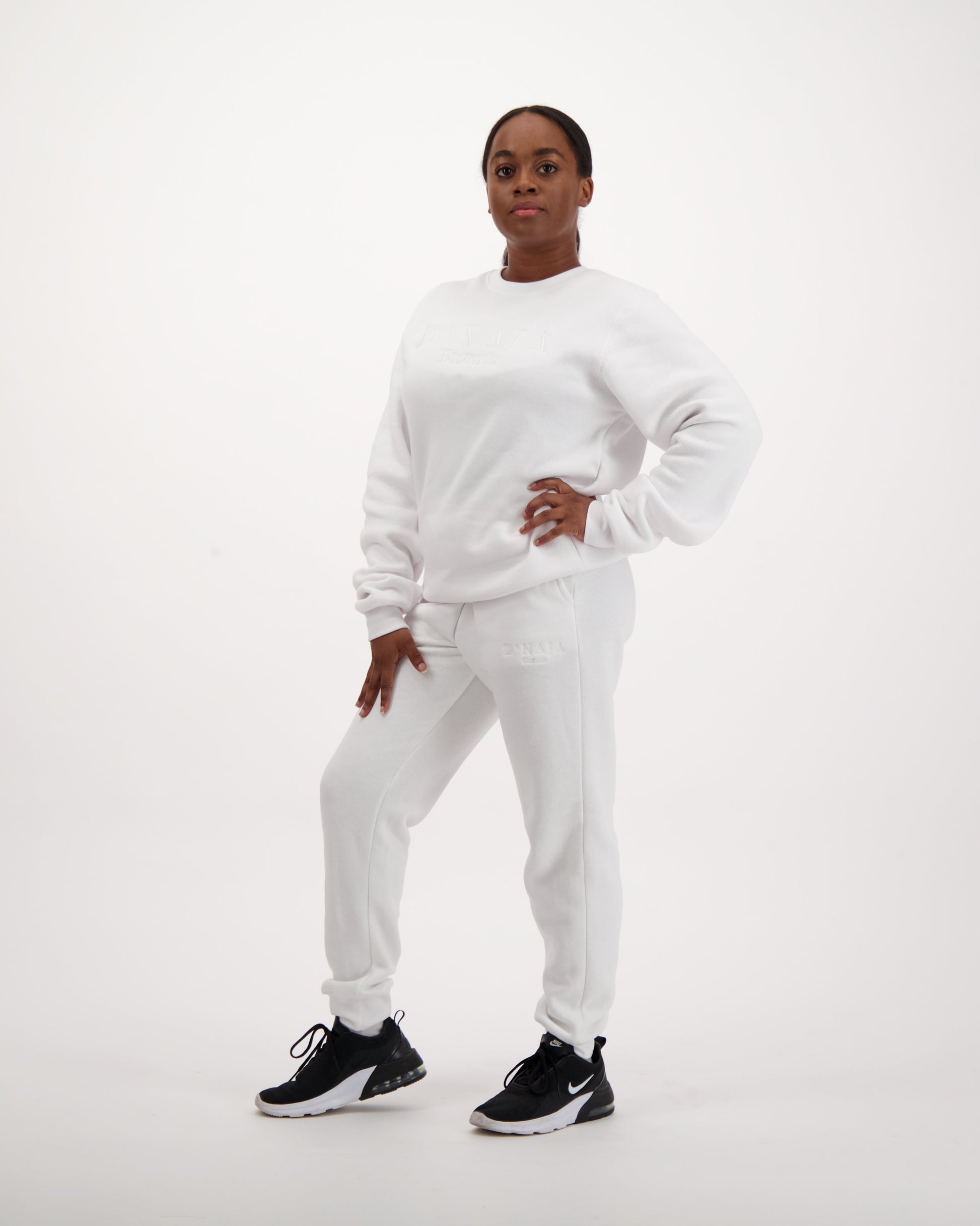 A woman wearing a white fleece crewneck sweatshirt with the D'naja logo prominently displayed, showcasing contemporary style and brand recognition